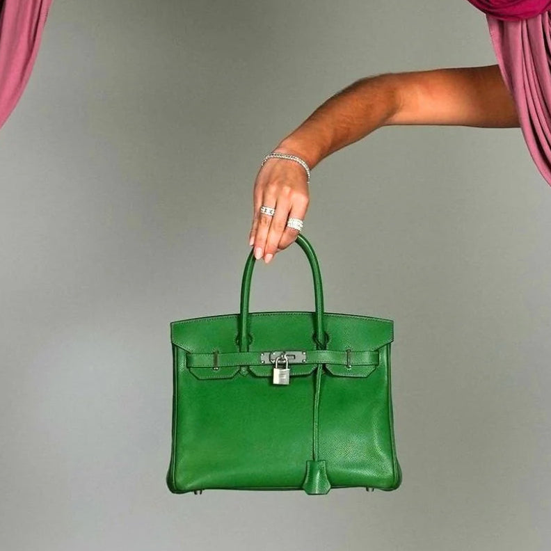 A bright green leather handbag with silver hardware is held by a person's arm, suspended above a beige pedestal between draped magenta curtains against a light gray background.
